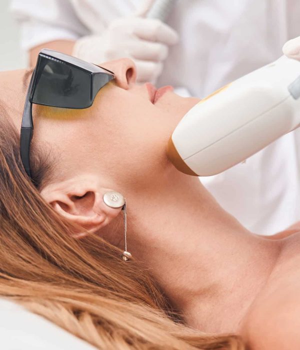 Calm woman on couch is wearing protective glasses while cosmetologist is using laser for face epilation